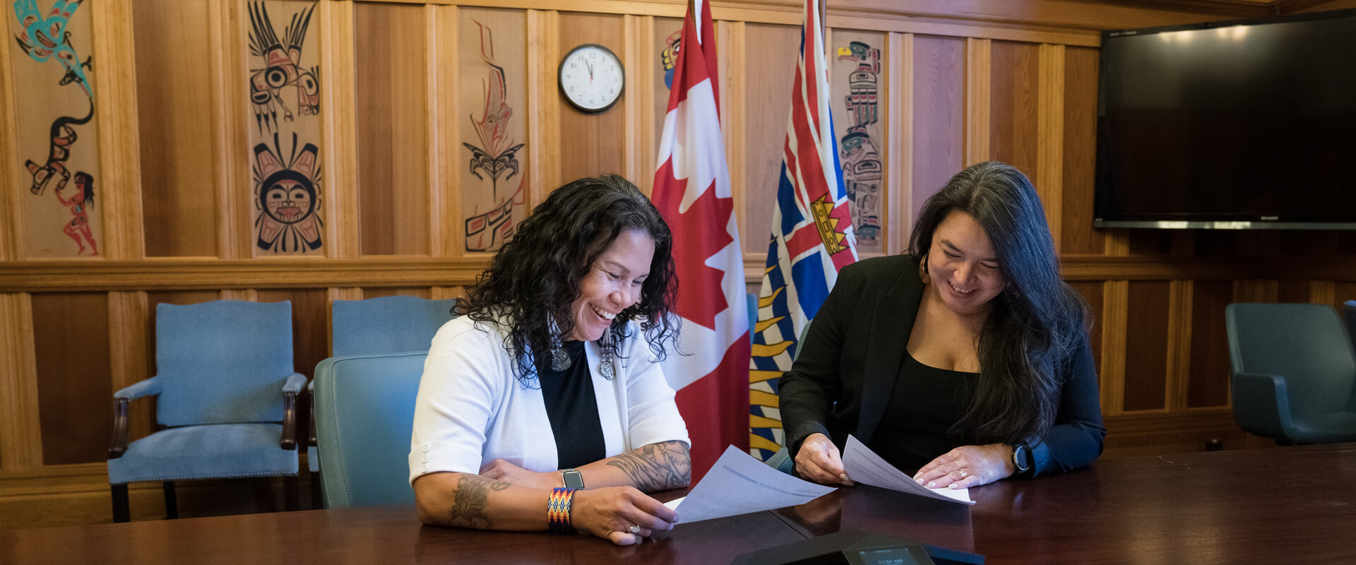 Jessica and Priscilla sitting together at a long table, smiling and laughing while handling papers. They sit in front of the BC and Canada flags, and the wood panelled wall has traditional artwork.