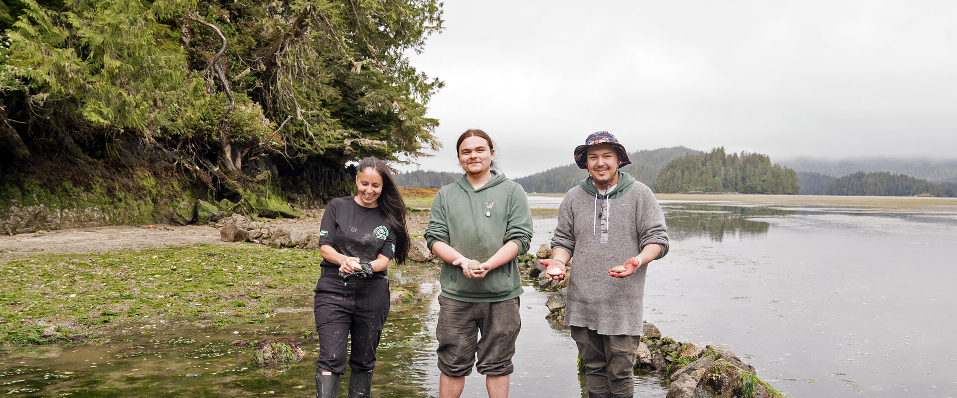 Three young people standing in the clam field, holding clams and smiling.