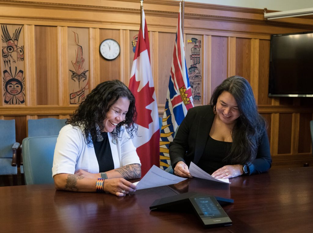Jessica and Priscilla sitting together at a long table, smiling and laughing while handling papers. They sit in front of the BC and Canada flags, and the wood panelled wall has traditional artwork.