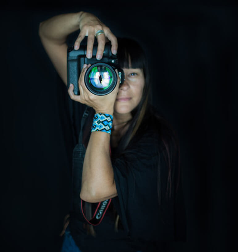 Artistic photo with black background of Melodie pointing a large camera lens at the viewer.
