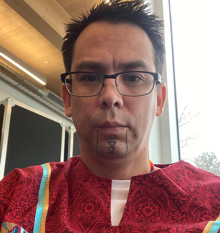 Sheldon wearing red shirt, glasses, with traditional Indigenous tattoos on his chin.