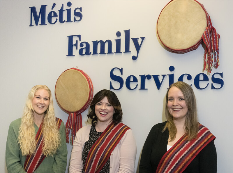 Three women smiling while wearing woven Métis sashes, standing under a wall with hanging drums and words "Métis Family Services."