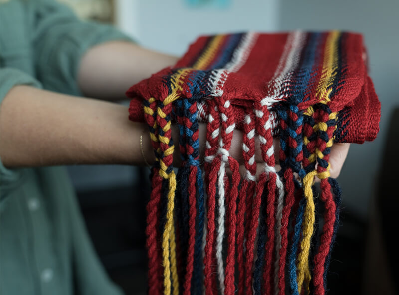 Folded woven sash in red, blue, yellow, white, and black rest on a person's outstretched arms.