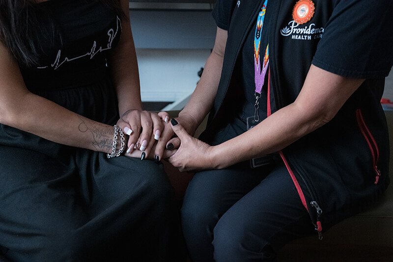 Close-up of Raquel and Ange's hands clasped on their laps. Both have decorated nails and wear dark clothes.