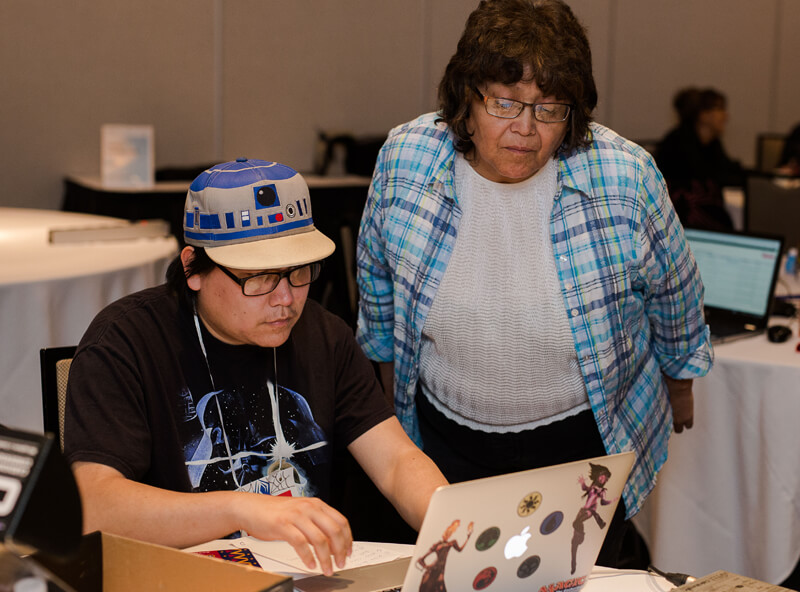 A man wearing R2D2 hat sits at a table with his laptop. A woman in a plaid blue shirt stands over him looking at the screen.