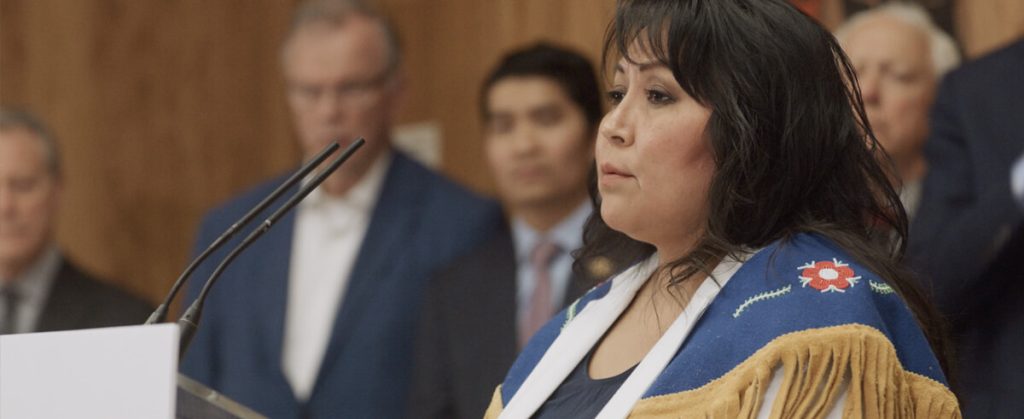 A woman wearing traditional Indigenous garment stands and speaks to government.