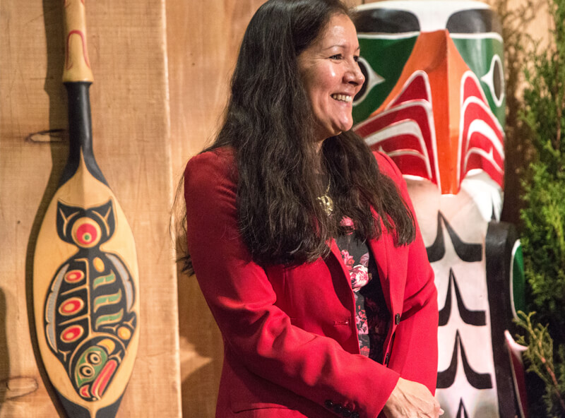 Deborah Jeffrey in a red blazer stands smiling in front of Indigenous artwork of a paddle with a fish painted on it and eagle statue.
