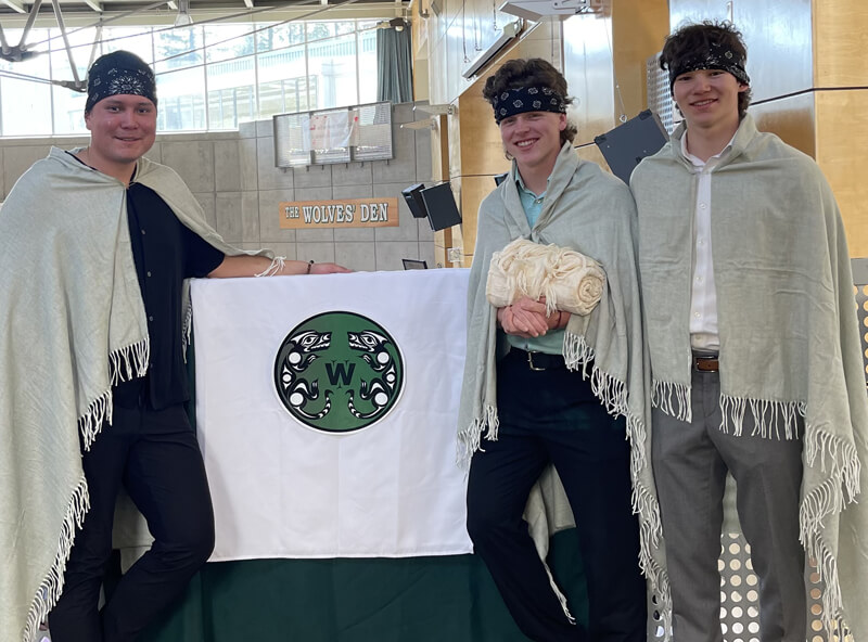 Three youth stand together, smiling at the camera, wearing tasseled shawls and bandanas next to a tapestry. A sign with "The Wolves' Den" written on it is seen in the background.