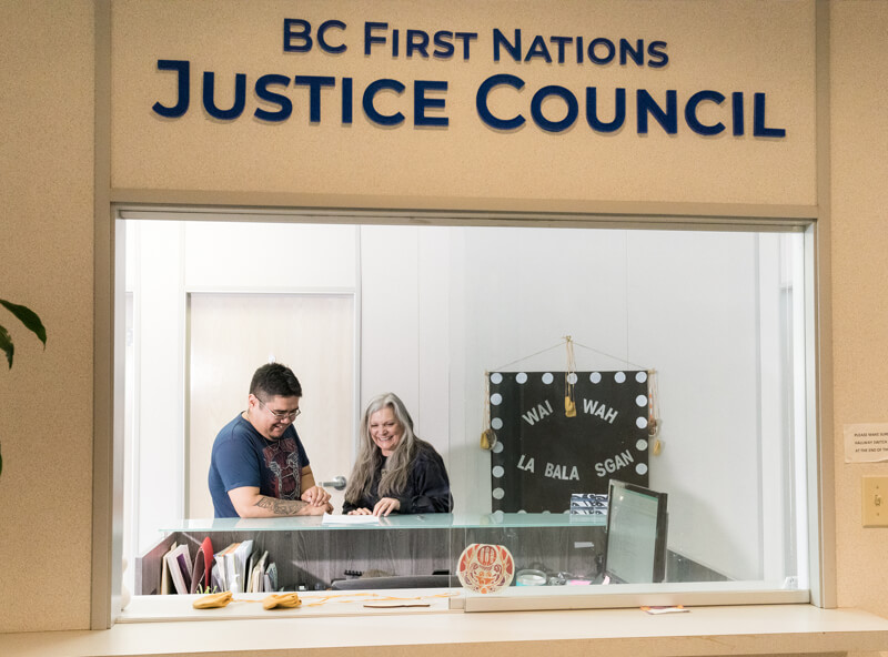Michael and a woman with long grey hair are seen at a front desk, smiling together, at the BC First Nations Justice Council.