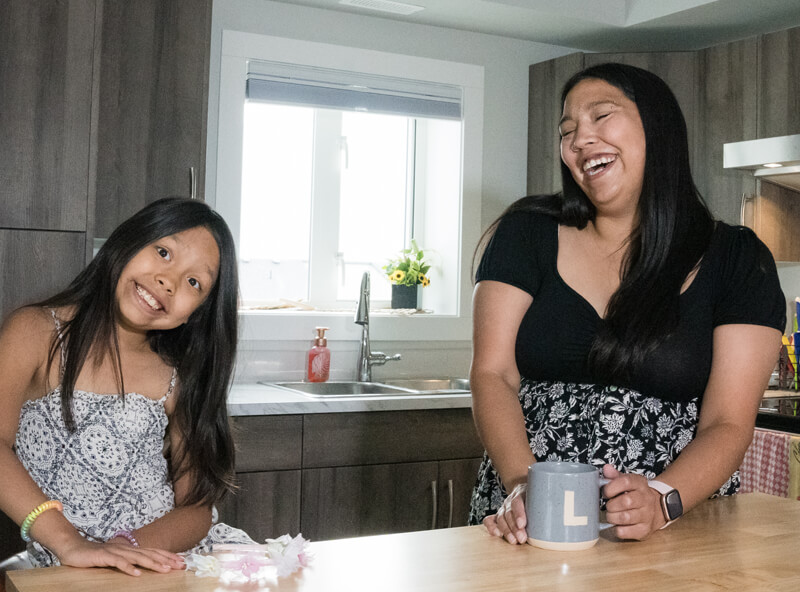 Lauren Marchand laughs in her kitchen as her daughter makes a funny face at the camera.