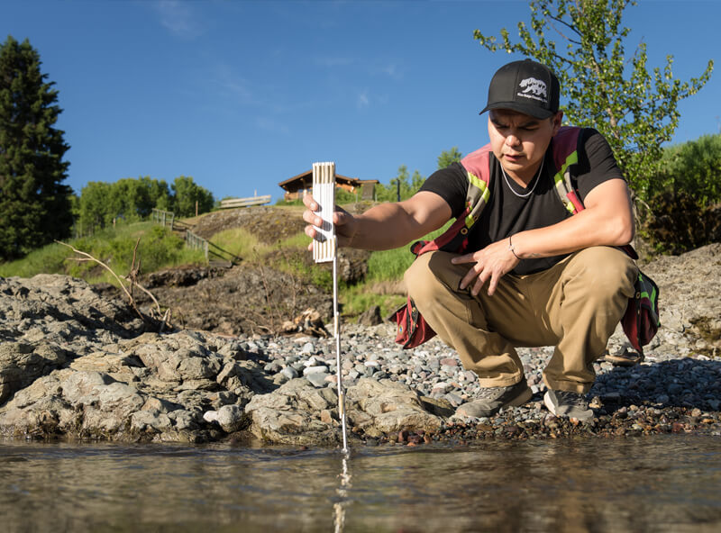 Dallas Nikal in hi-vis vest and black baseball cap crouched next to water, holding device in the water to take environmental data.