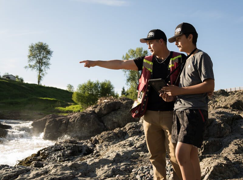 Dallas Nikal points out across rushing water, guiding the attention of a young adult standing next to him.