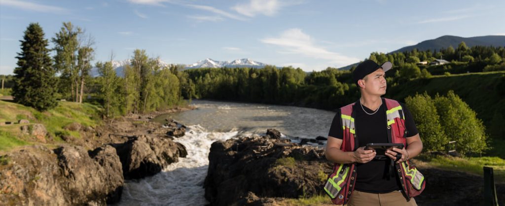 Dallas Nikal in hi-vis vest and black cap looking to the right. Behind him, a body of water surrounded by forest and mountain range is in the distance.